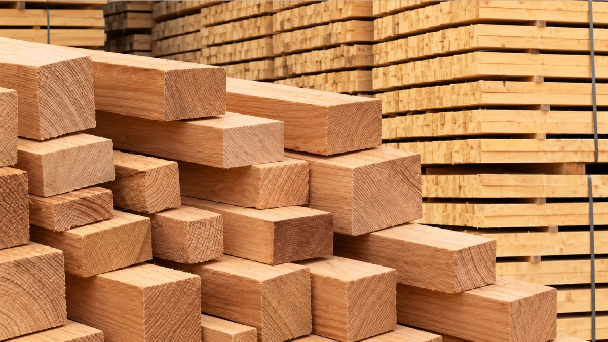 Gestapelte Holzbalken mit rechteckigen Querschnitten im Vordergrund. An den Stirnseiten sind Jahresringe und Holzstrukturen sichtbar. Dahinter lagern Bretter in gleichmäßigen Reihen. Warme, goldbraune Farbtöne prägen das Bild.